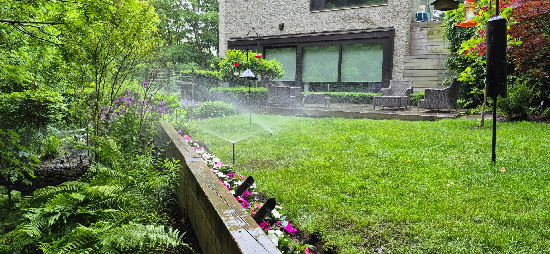 Pop-up sprinklers watering a backyard lawn beside garden beds and a patio after spring start-up