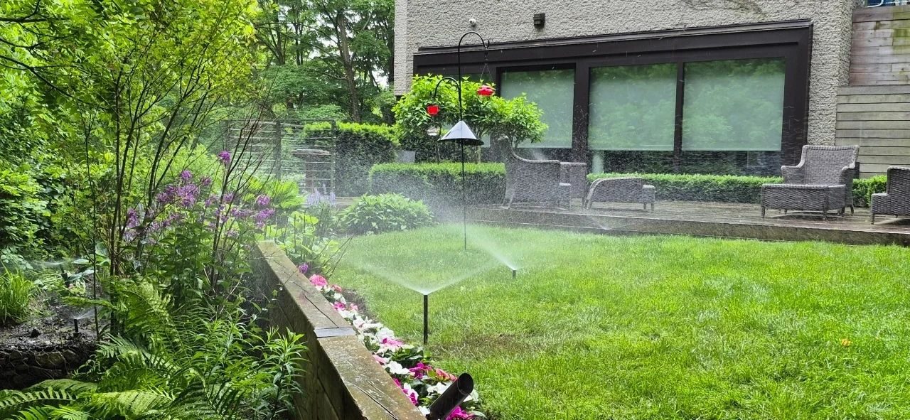 Pop-up sprinklers watering a backyard lawn beside a raised flower bed and a wooden deck